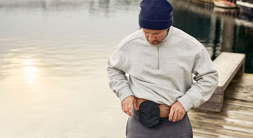 Mann med lue står på en bro ved havet. En svart stomipose er synlig på magen.
